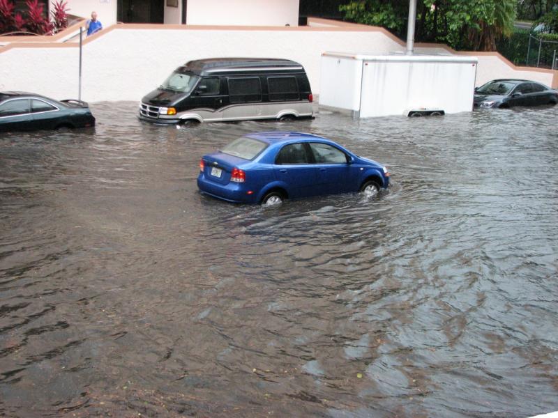 Photograph of a flooded street with a blue compact car in the middle of the road, with water up to the bottom of the chassis.