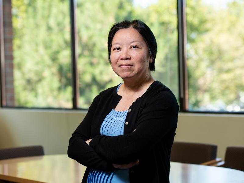 A woman with short black hair in black cardigan and periwinkle shirt smiling in front of a window in a conference room