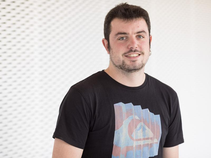 Smiling man with light skin and dark hair wearing a black t-shirt on a lightly patterned white and gray background