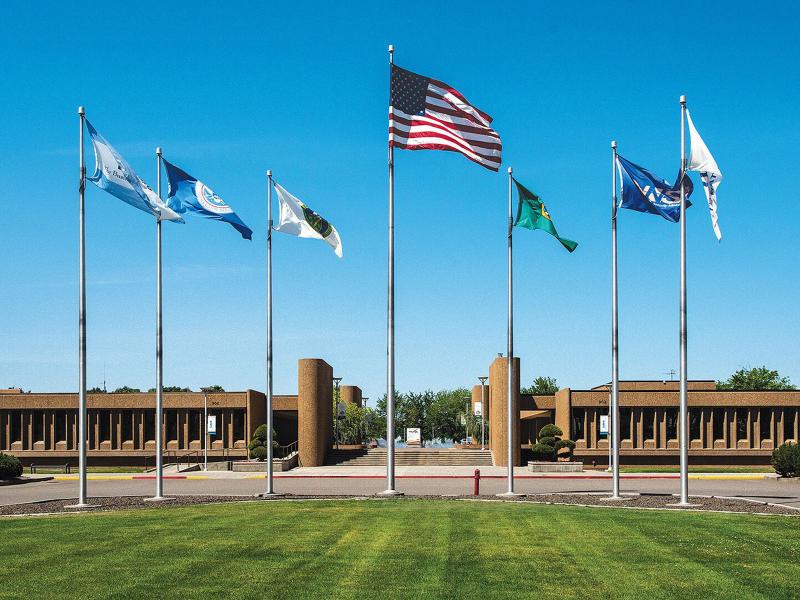 A collection of poles flying various flags on a grass field in front of a collection of brown stone buildings