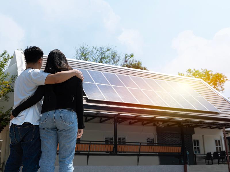 Photo shows two people with their arms around each other looking at their house, which has rooftop solar panels installed.