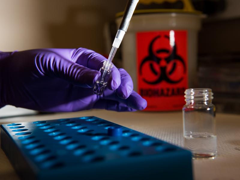 A gloved hand prepares a protein sample with a bio-hazard disposal container in the background.