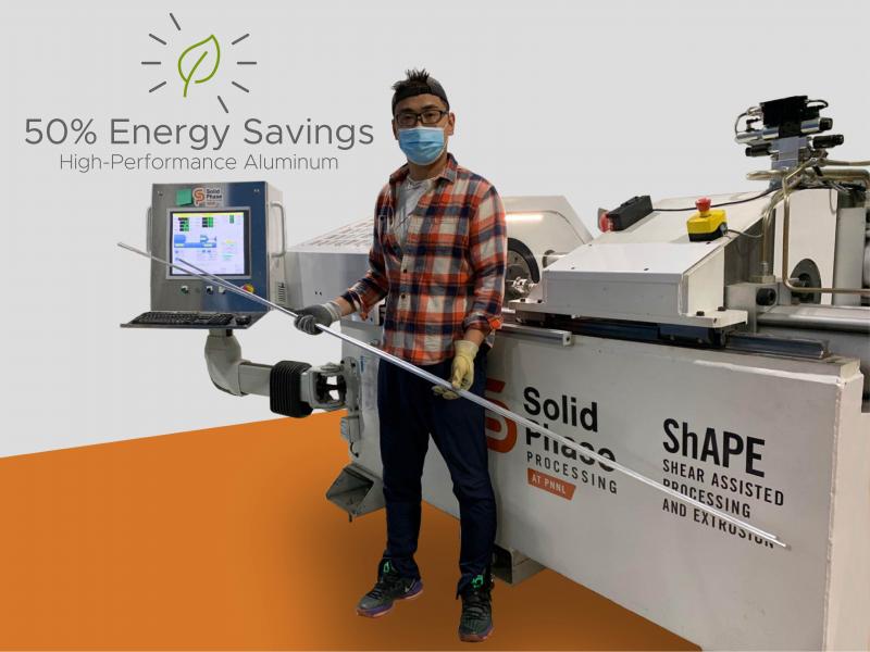 A researcher stands in front of the ShAPE machine holding a piece of aluminum alloy 7075, with the words "50% Energy Savings High-Performance Aluminum Alloy" in the background.