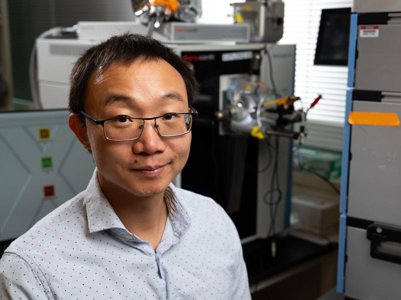 Photograph shows Tong Zhang on the left, looking into the camera, with laboratory equipment behind him and to the right.