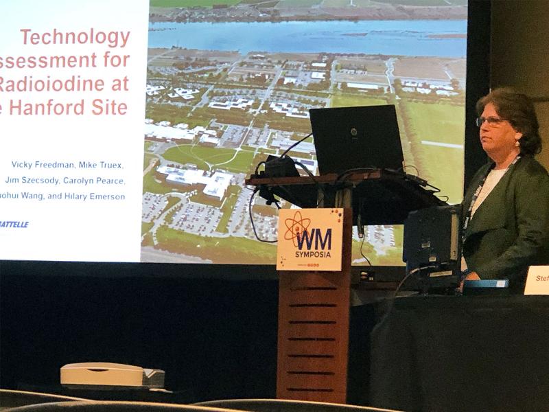 Vicky Freedman, a computational subsurface hydrologist, stands at a podium next to a big screen showing the title slide of her presentation