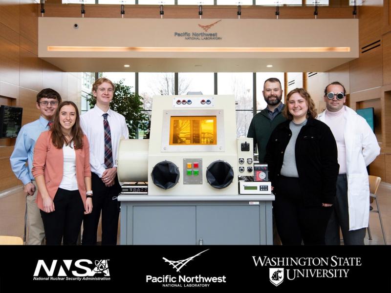 Photo of six people gathered around a machine located inside a PNNL building. Beneath the photo is a banner, featuring the National Nuclear Security Administration (NNSA,) Pacific Northwest National Laboratory (PNNL,) and Washington State University (WSU) names and logos.