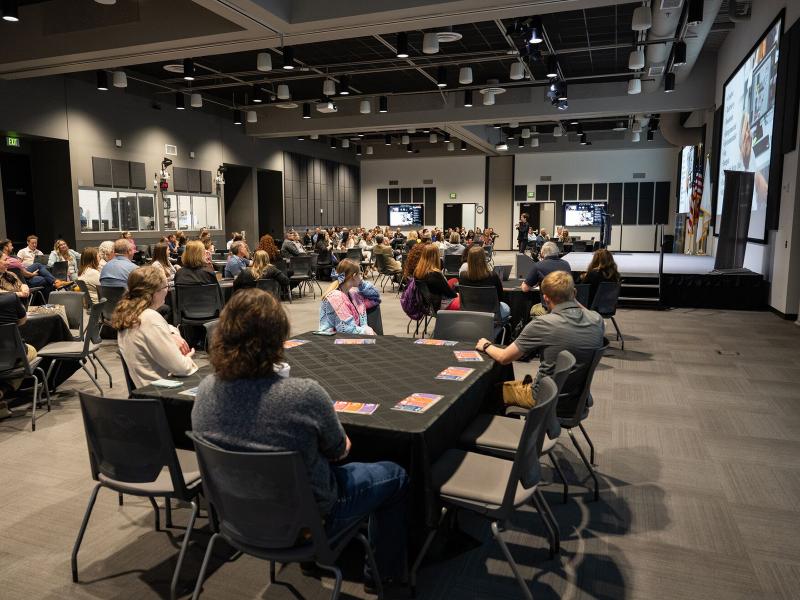 A large open hall with people seated in groups at round tables listening to a speaker standing on an elevated platform in front of a large screen