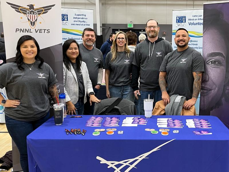 Group photo of PNNL staff members smiling at the camera from behind a career fair table. In the background is a stand-up banner that says, "PNNL Vets."