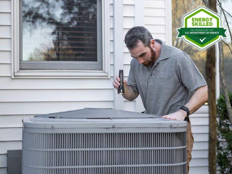 a man stands over an air conditioner, looking at it with a flashlight. In the corner there's a green and white DOE Energy Skilled badge