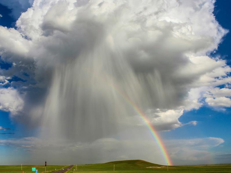 a very tall, large storm cloud with rain and a a rainbow over the countryside 