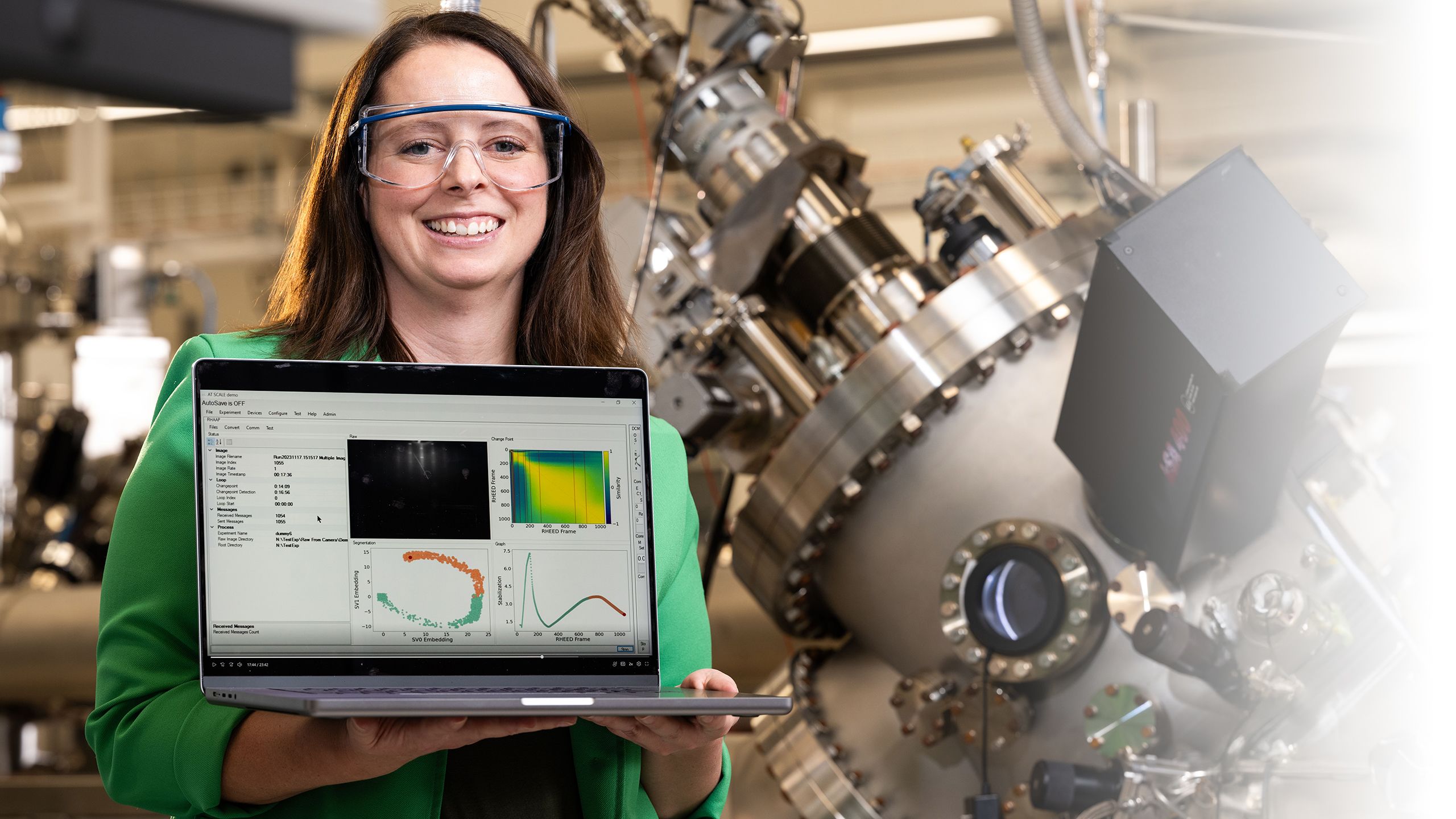 a smiling researcher wearing safety glasses and holding a laptop turned toward the camera to show graphs and charts on the screen