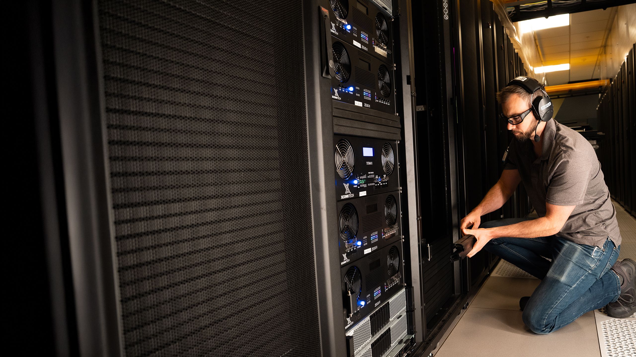 a technician wearing safety goggles and ear protection works on a large bank of servers