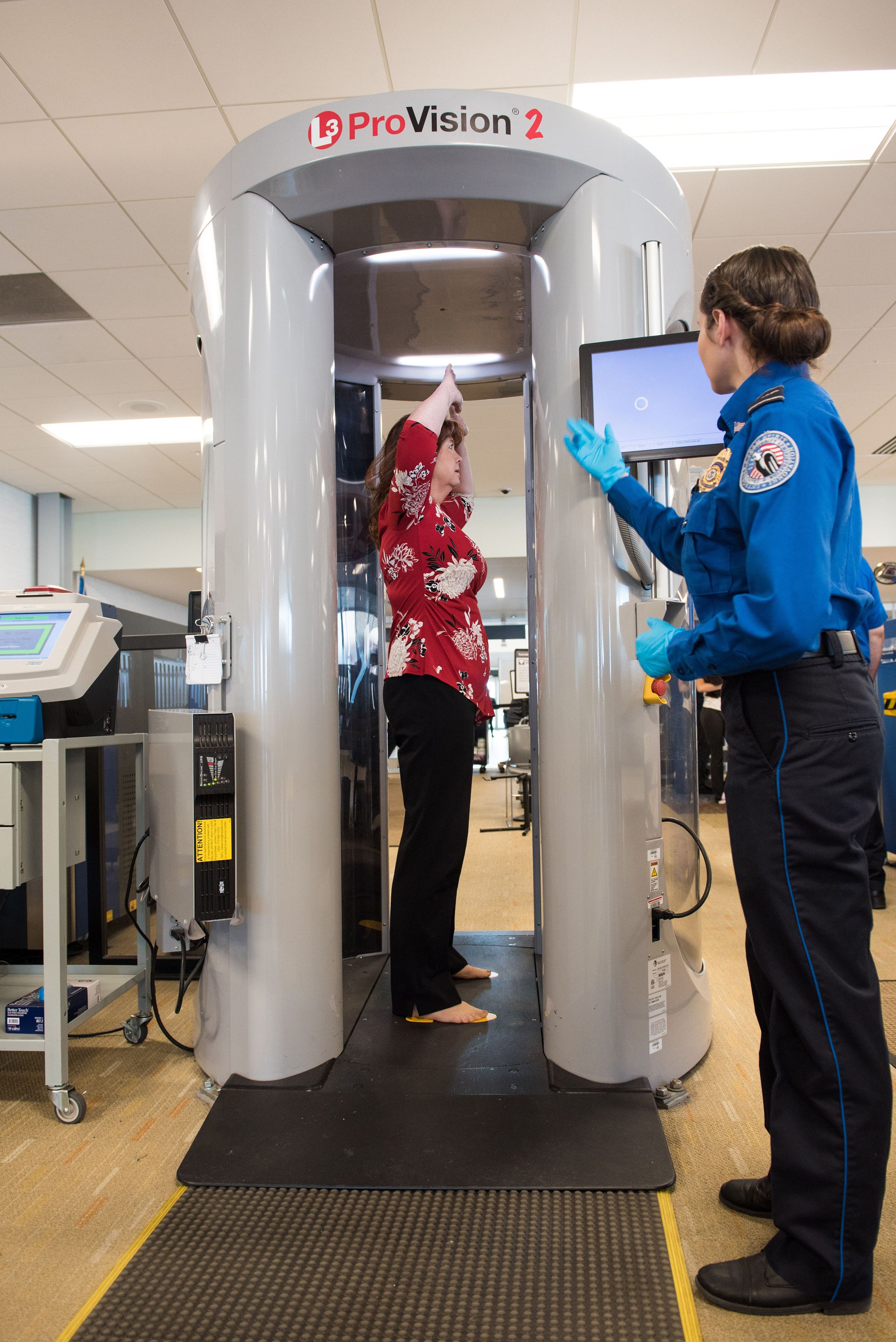 A passenger stands in a 360-degree airport security scanner while a security person watches