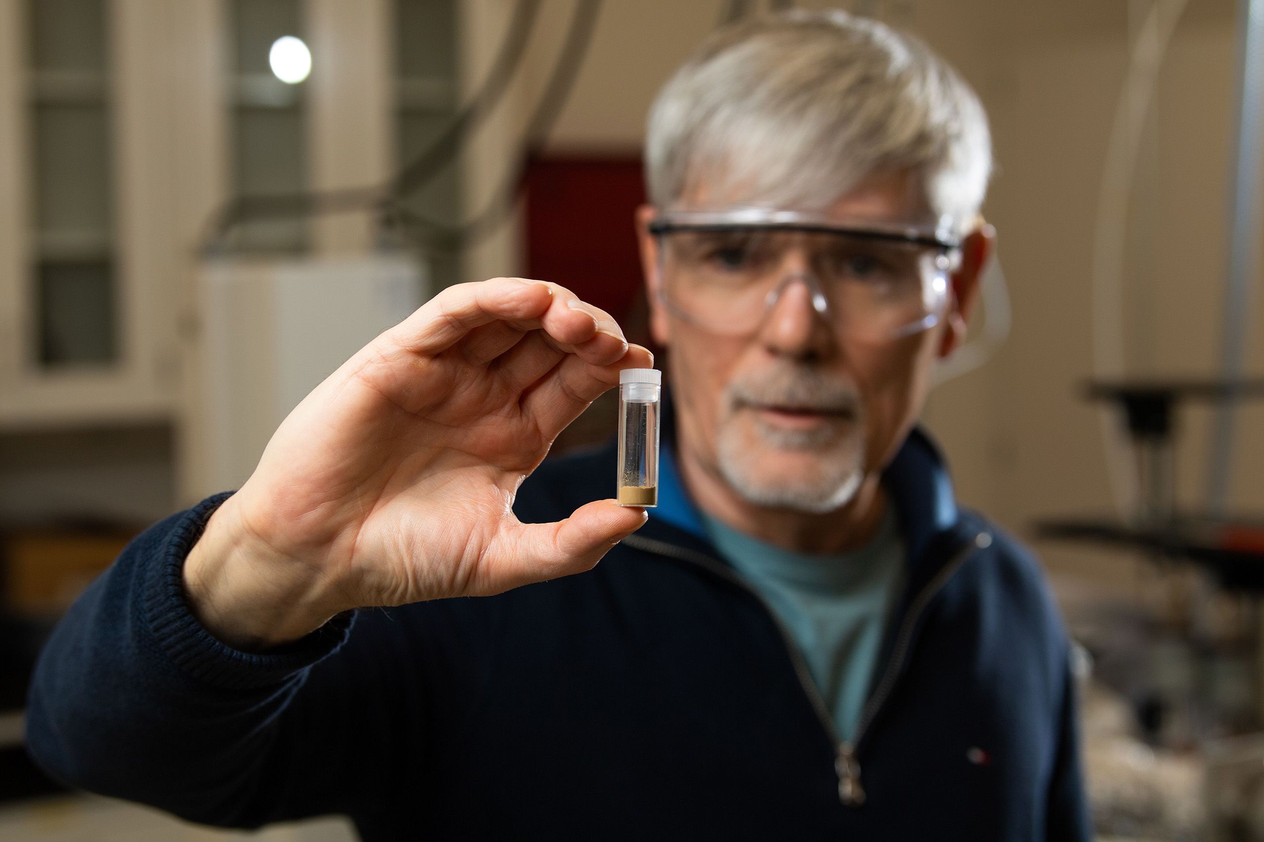 a researcher in safety glasses holds up a glass vial with a small amount of material in the bottom