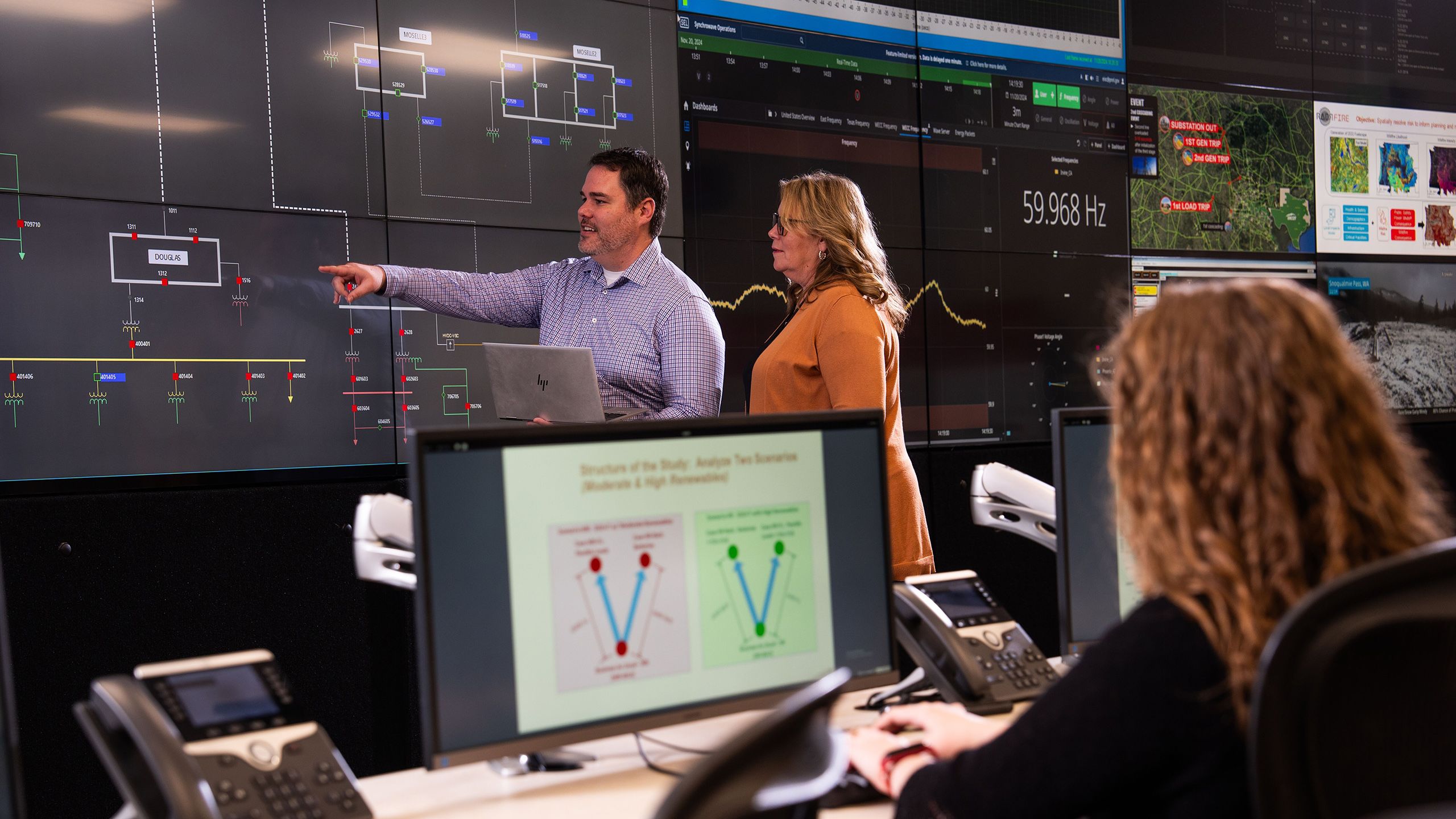 two researchers review what is displayed on a large video grid wall while another researcher in the foreground works on a computer