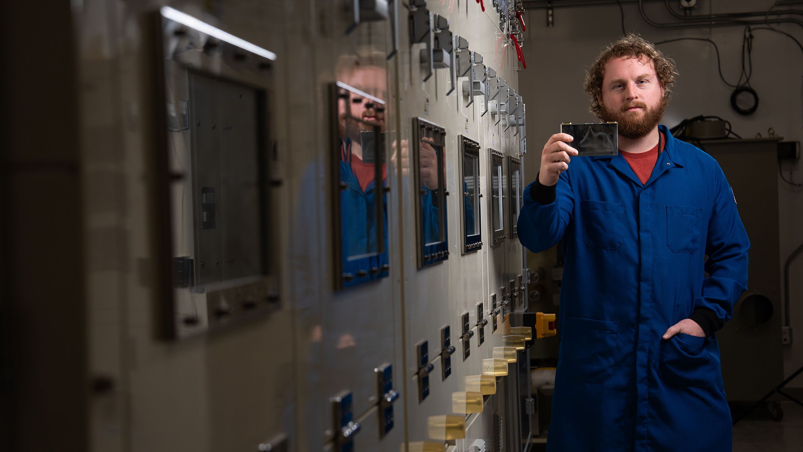 A researcher in a jumpsuit holds a rectangular, reflective black object in a lab hallway